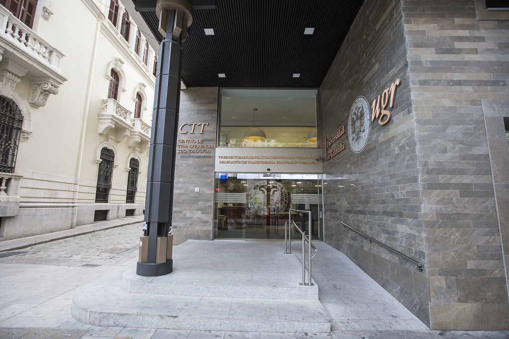 Aparece una imagen que muestra la entrada principal del Centro de Transferencia Tecnológica (CTT) de la Universidad de Granada. En la fachada se distinguen claramente los logotipos y textos institucionales de la UGR. La arquitectura combina elementos modernos con el entorno clásico de los edificios adyacentes.