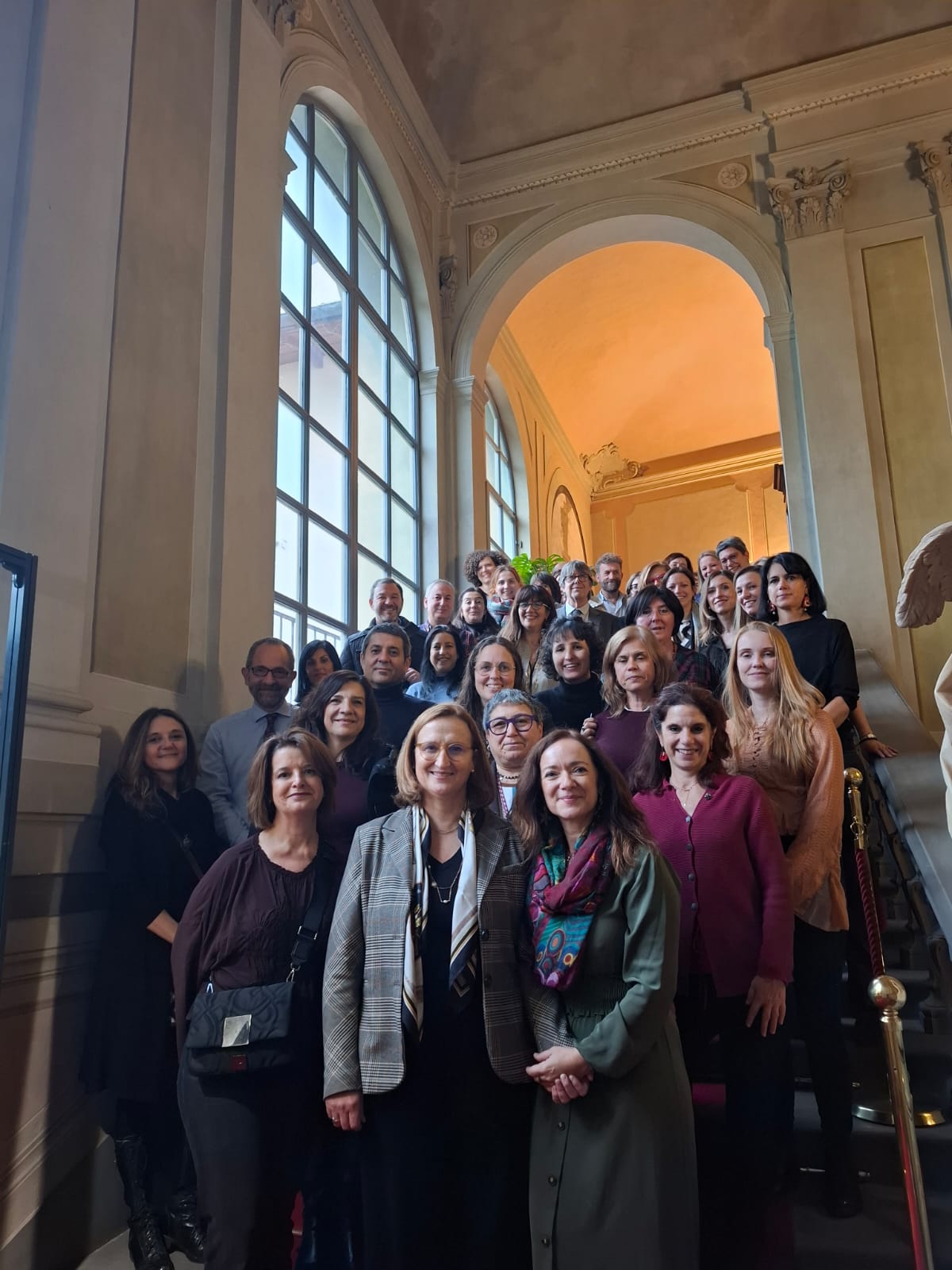 Aparece una imagen de grupo tomada en una majestuosa escalera interior con grandes ventanales y luz natural. Las personas están alineadas en varios niveles, sonriendo, en un entorno elegante de arquitectura clásica. Los colores predominantes son beige, marrón, gris y toques de burdeos y verde.
